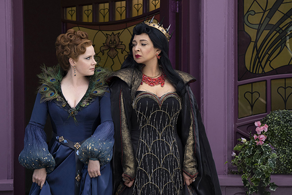 Two women in ornate fantasy gowns and a crown stand before a purple building with stained glass.