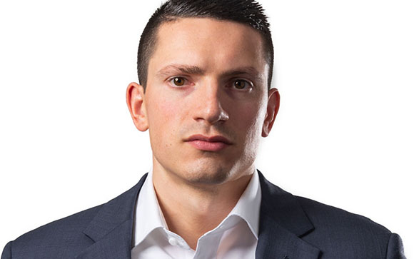 A man with short dark hair wearing a white shirt and dark suit jacket against a white background.