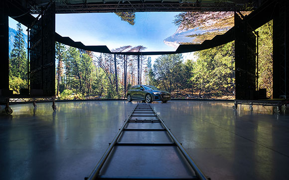A silver car sits on a studio floor in front of a large screen displaying a lush forest and blue sky.