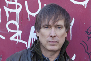 A man with brown hair and a serious expression stands before a red wall covered in white graffiti.