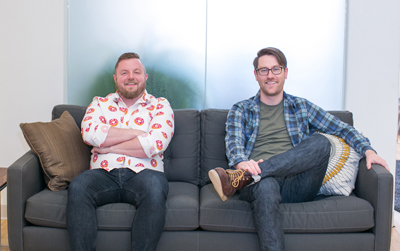 Two men smiling while sitting on a dark gray couch against a frosted glass background.