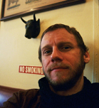 A man with short hair and a beard looking toward the camera against a cream-colored wall.