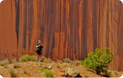A person in hiking gear stands in front of a towering, vertical red sandstone cliff face.