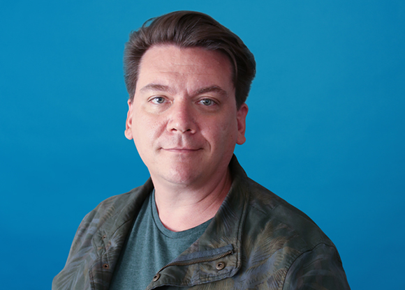 A man with brown hair wearing a green t-shirt and a camouflage jacket against a solid blue background.