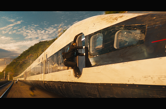 A person in dark clothing clings to the exterior of a fast-moving white and black train.