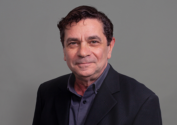 A middle-aged man with dark wavy hair wearing a dark blazer and a grey button-down shirt against a grey background.