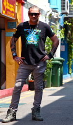 A man wearing sunglasses, a black graphic t-shirt, grey pants, and boots poses on a sunny street.