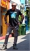 A man in a black graphic t-shirt and grey trousers poses with hands on hips on a sunny street.