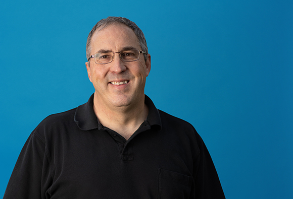 A smiling man with glasses and short hair wearing a black polo shirt against a solid blue background.