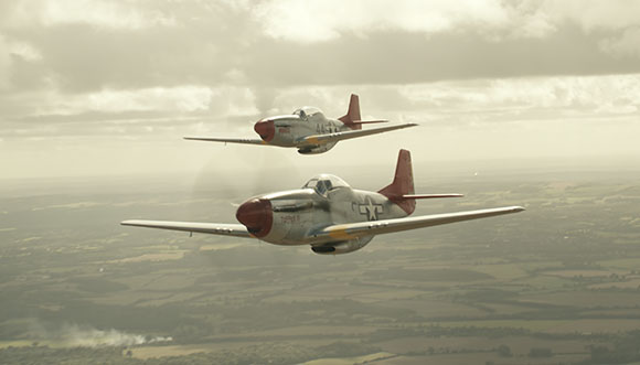 Two silver fighter planes with red noses flying in formation over a green countryside under a cloudy sky.