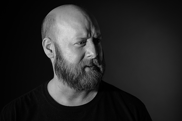 Black and white studio portrait of a bald man with a full beard looking away from the camera.