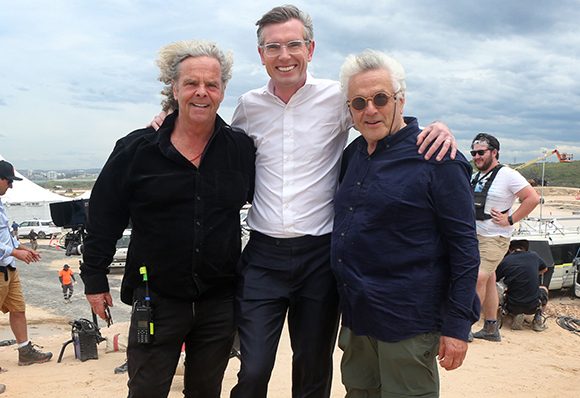 Three men smiling and posing together on a sandy outdoor set under a cloudy sky.