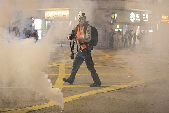 A photographer in an orange vest and gas mask walks through heavy white smoke on a city street.