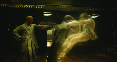A person in a light green robe stands facing several blurred, fast-moving figures in a dimly lit room.