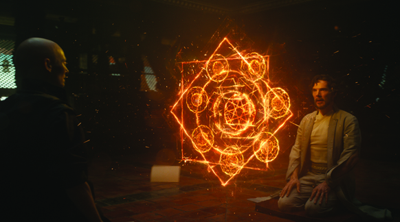 A man sits in meditation facing a large, glowing orange magical mandala in a dark room.