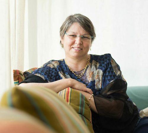 A woman with short grey hair and glasses smiles while leaning on a colorful striped cushion.