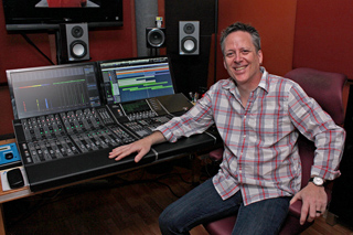 A man in a plaid shirt sits in a recording studio next to a large digital mixing console and monitors.