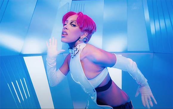 A person with short pink hair and white gloves poses against a blue, geometric background.