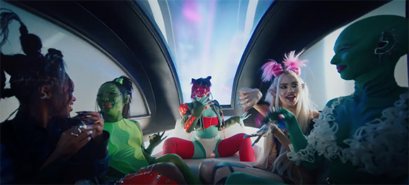 Five people in colorful, otherworldly costumes and makeup sit together inside a futuristic vehicle.