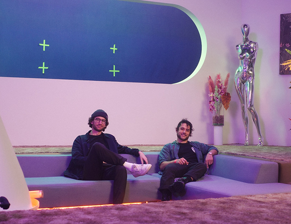 Two men sit on a long purple sofa in a room with a large blue wall graphic and a silver statue.