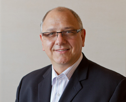 A middle-aged man with glasses and a suit smiles at the camera against a neutral background.
