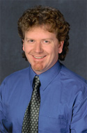A man with curly brown hair, wearing a blue button-down shirt and a patterned tie, smiling against a dark background.