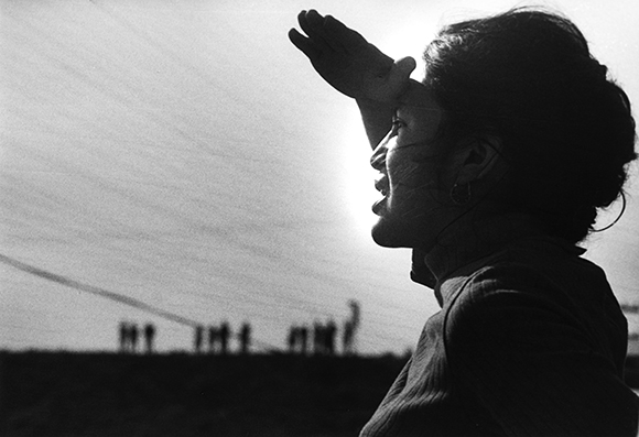 Black and white silhouette of a woman shielding her eyes with her hand against a bright sky.