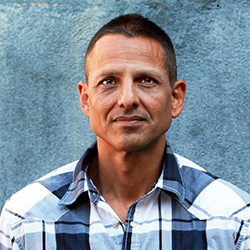 A man with short dark hair wearing a blue and white plaid shirt against a textured blue wall.