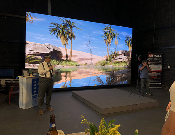 A man in a uniform holds papers in front of a massive screen showing a desert oasis with palm trees.