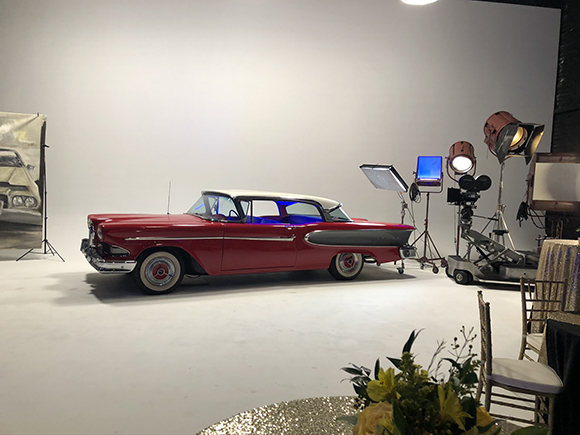 A red vintage sedan with a white roof parked in a white studio surrounded by professional lighting equipment.