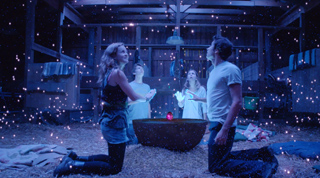 Four people kneel in a dark barn, looking up at glowing particles floating in the air.