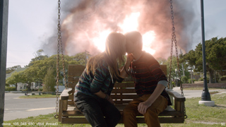A couple kisses on a wooden porch swing as a massive explosion erupts behind them.