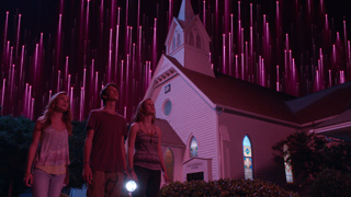 Three young people stand in front of a white church looking up at pink light streaks in a dark sky.