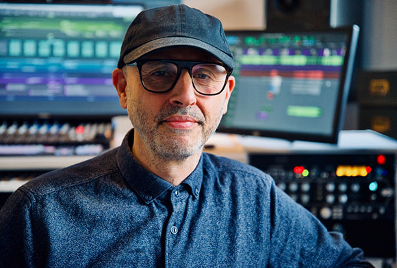 A man with glasses and a dark cap sits in front of computer monitors and audio equipment.