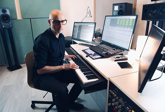 A bald man with glasses playing a digital piano at a desk with computer monitors and studio speakers.