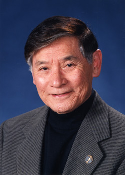 Headshot of an elderly man with dark hair, wearing a grey blazer and black turtleneck against a blue background.