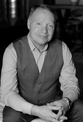 A man wearing a button-down shirt and a dark vest sits with his hands clasped.
