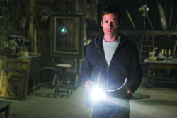 A man in a dark, cluttered room holds a bright flashlight, illuminating the space in front of him.