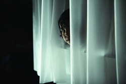 A person's face partially visible while peeking through vertical white curtains in a dark setting.