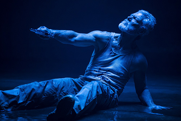 A man in a white tank top and pants sits on a dark floor, holding a handgun out to the side.