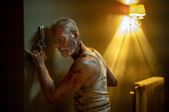 A bloodied man in a tank top holds a handgun against a wall under warm light.