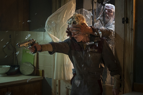 A person in a jumpsuit aims a handgun while an older man reaches through a broken window to cover their eyes.