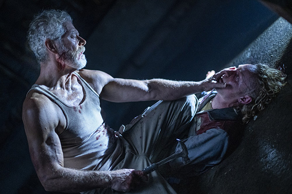 A man in a white tank top grips the chin of a man lying against a dark wall.
