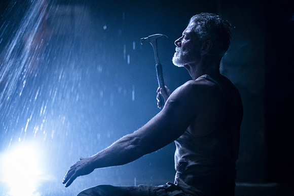 A man in a tank top sits in the dark, holding a hammer while rain falls around him.