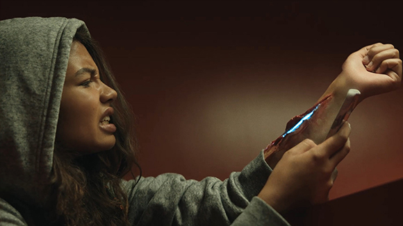 A person in a grey hoodie looks in pain at a glowing, metallic device embedded in their forearm.