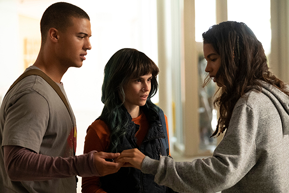 Three young people stand in a bright hallway, engaged in a serious conversation.