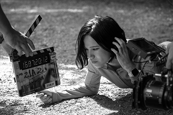 A woman leans close to a professional camera on a tripod while holding a clapperboard in a black and white photo.