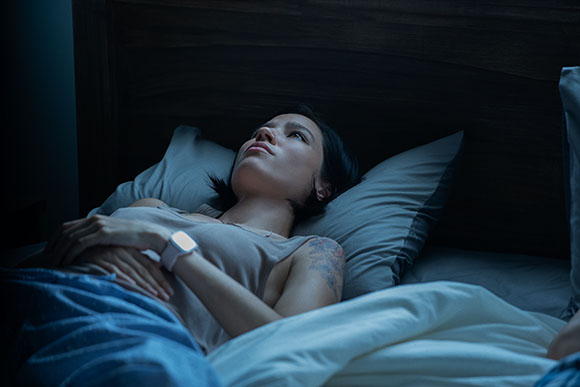 A woman with short dark hair lies awake in a dark bedroom, staring upward while resting on pillows.