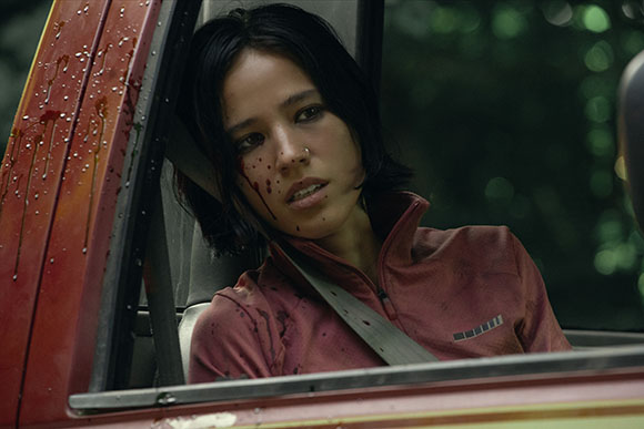 A young woman with dark hair and blood on her face sits in a red vehicle wearing a seatbelt.