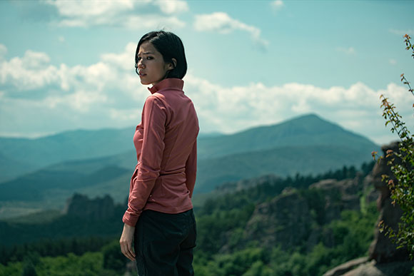 A woman in a pink shirt looks back while standing in front of a vast mountain range under a cloudy sky.
